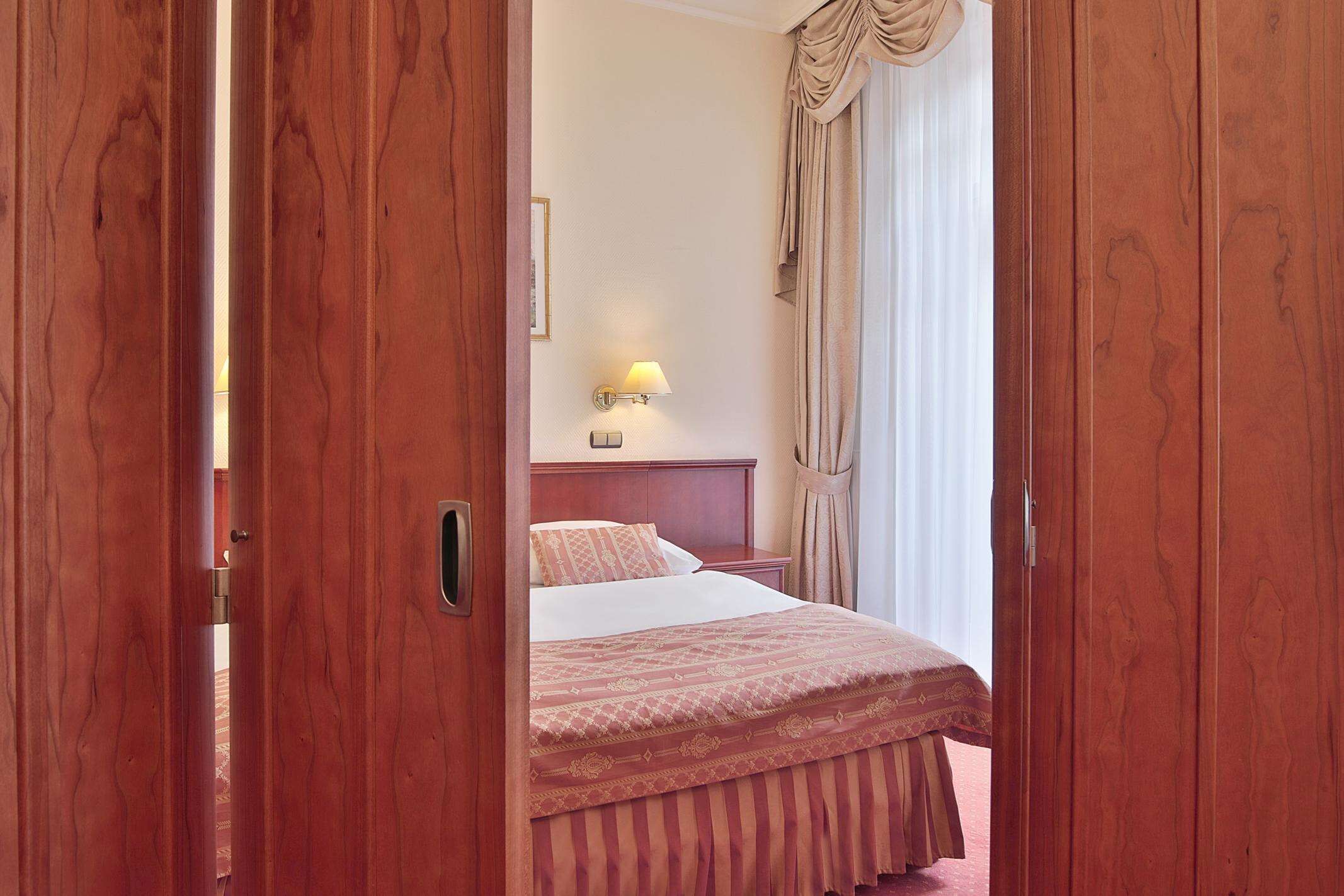 Una habitación de hotel con una puerta de madera, una cama con colcha estampada, una mesita de noche, una lámpara y una ventana con cortinas.