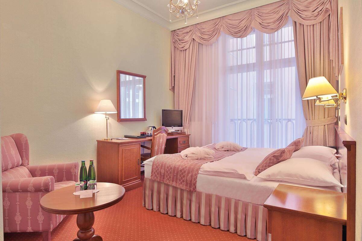 A hotel room with a bed, a desk with a computer, a chair, a small table with bottles, and a window with curtains.