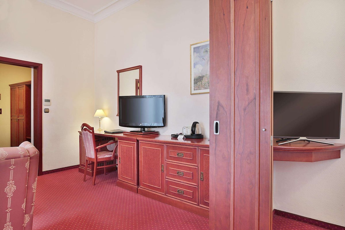 A hotel room with a television, a desk with a lamp, a chair, and a wardrobe.