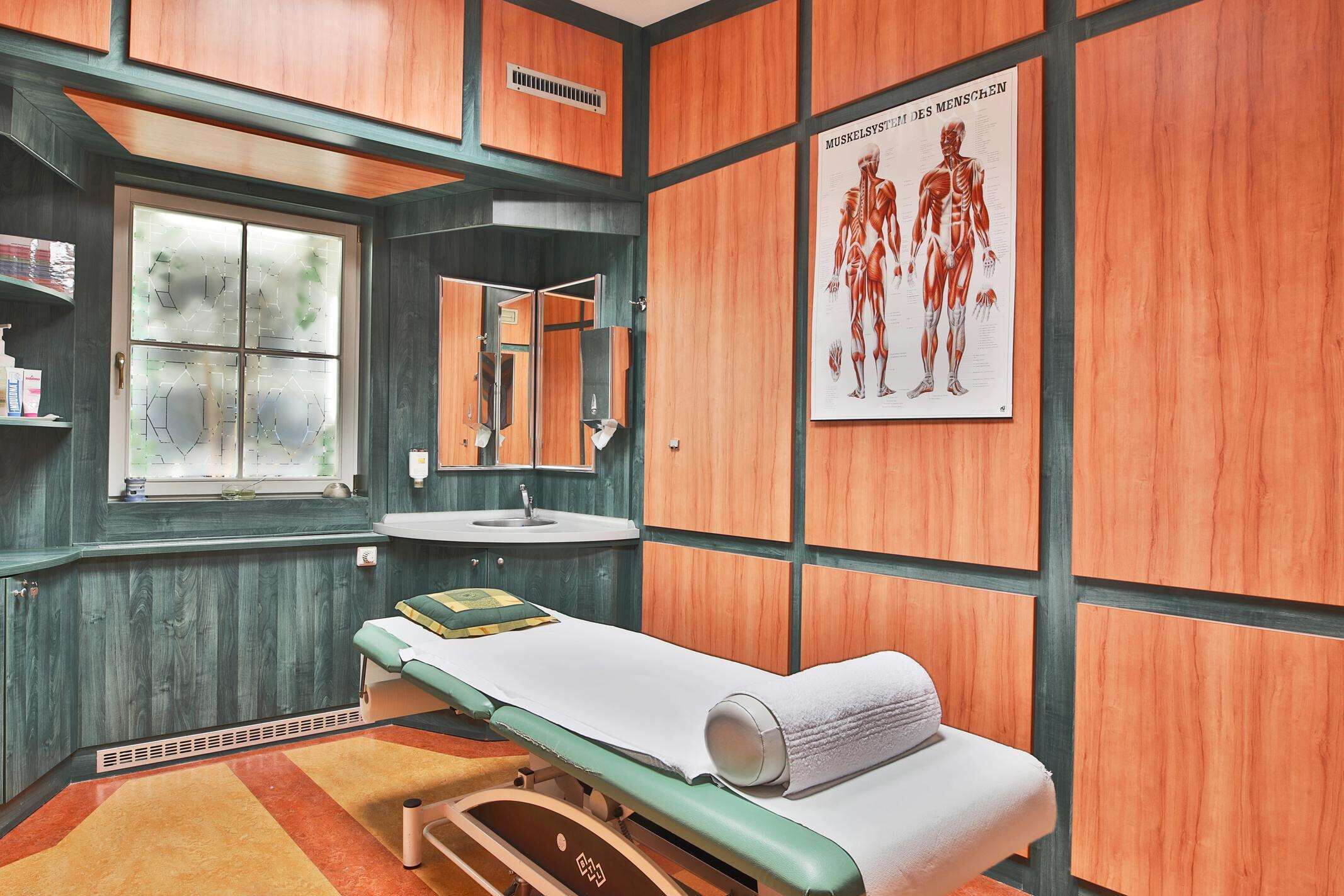 Un consultorio médico con pared de madera, una cama individual, un lavamanos y un cartel de anatomía humana.