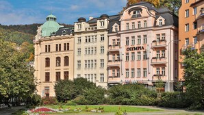 A historic building with the name "OLYMPIA" on the facade, surrounded by lush gardens and a clear sky.