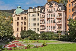 Un edificio histórico con el nombre "OLYMPIA" en la fachada, rodeado de jardines exuberantes y un cielo despejado.