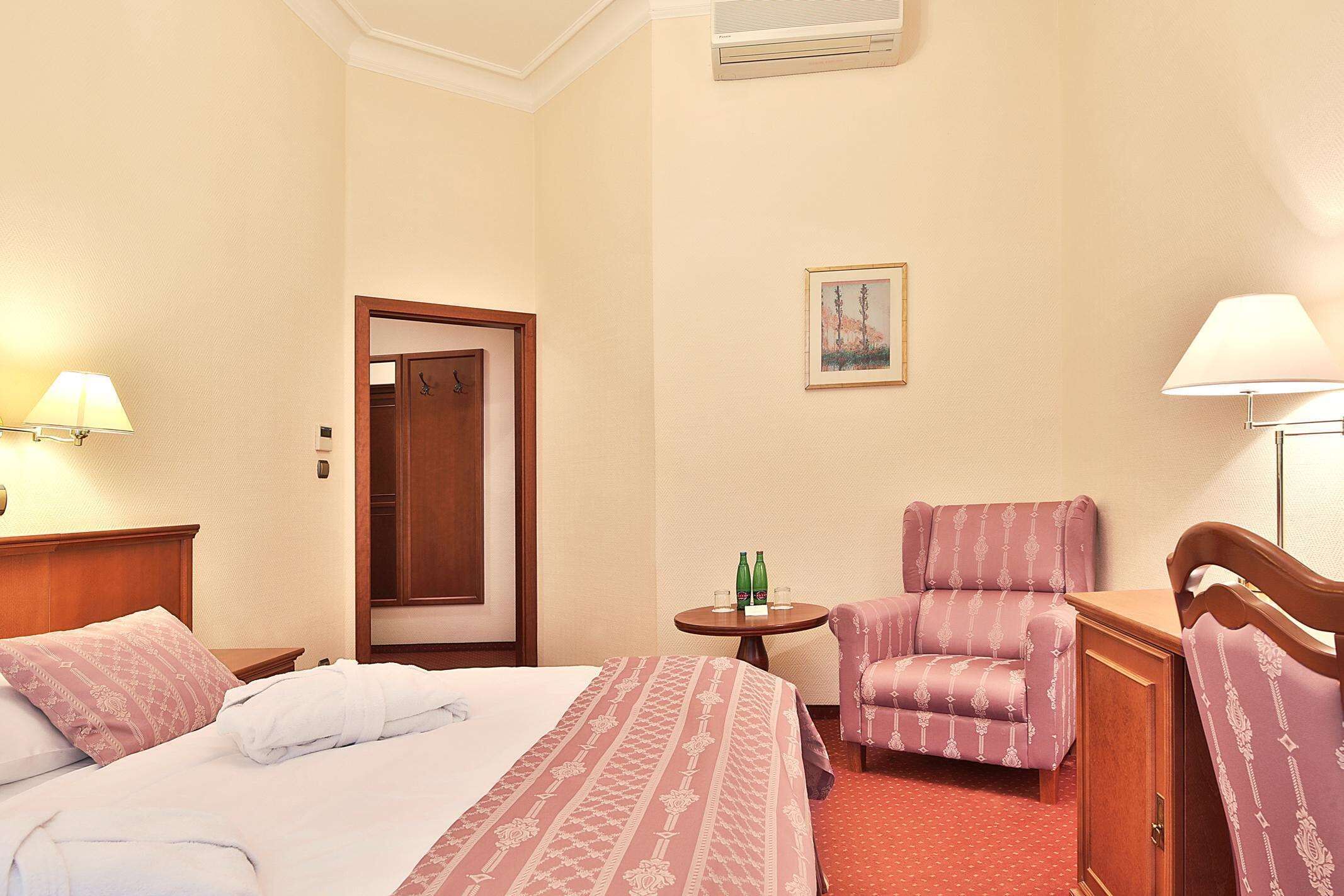 Habitación de hotel con cama, una silla, una mesita con botellas y un cuadro en la pared.