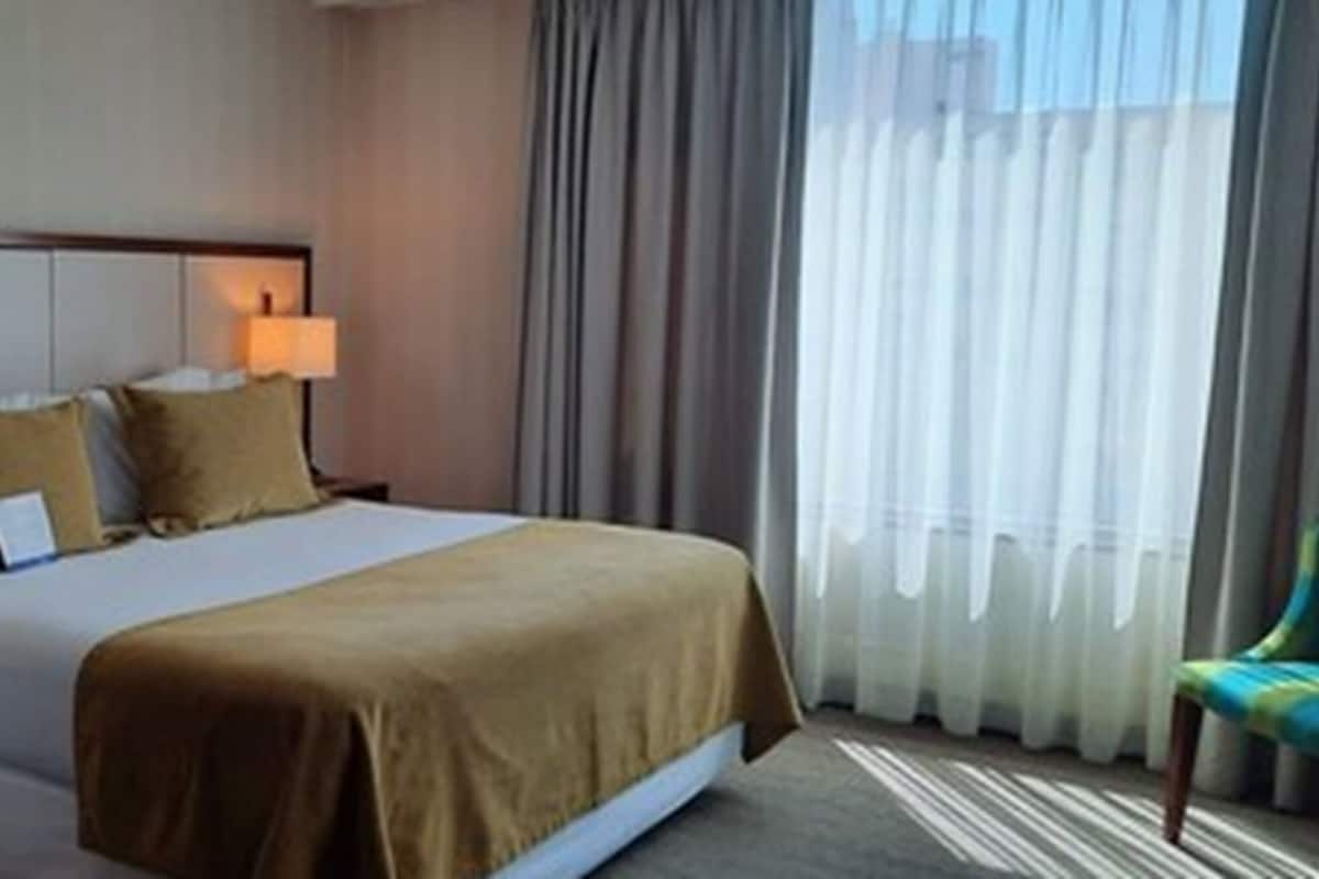 Een hotelkamer met een bed, twee bedlampen, een stoel en een raam met gordijnen.