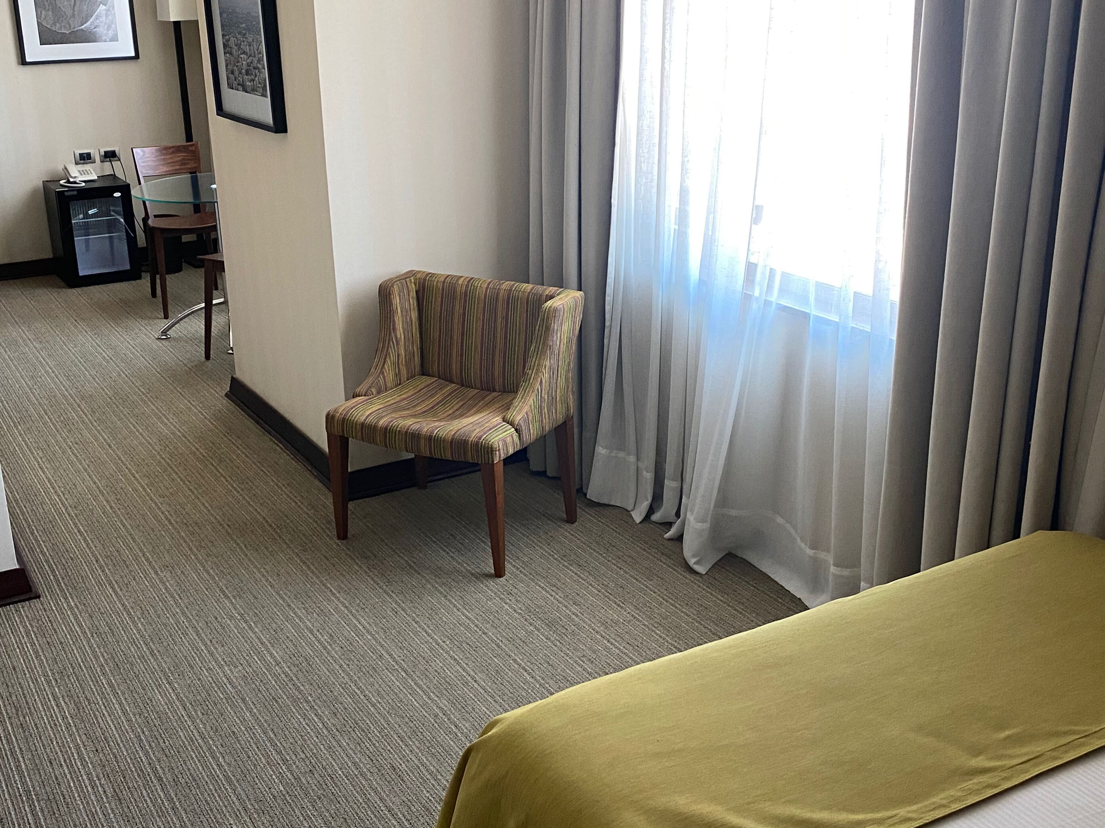 A hotel room with a bed, a striped armchair, a desk with a computer, and a window with sheer curtains.