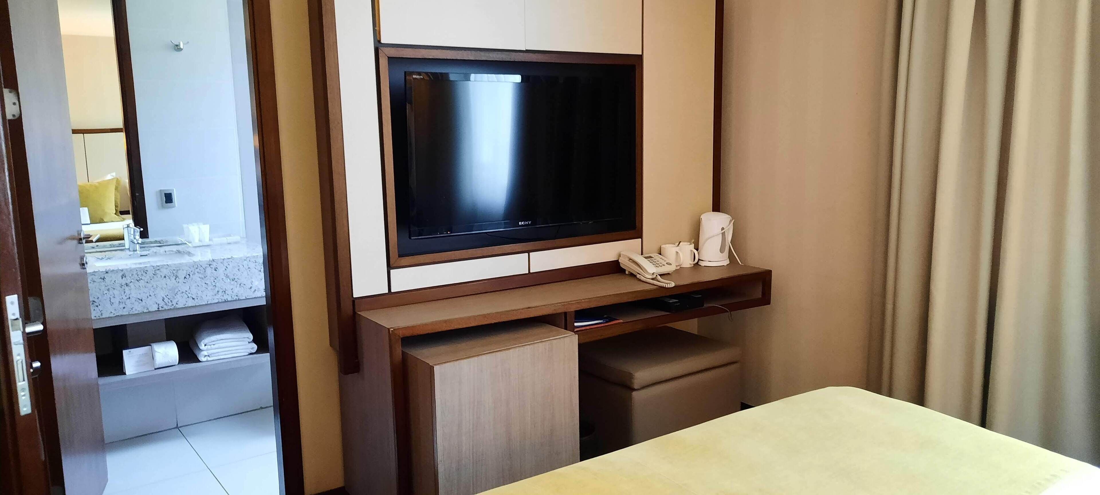 Una camera d'albergo con TV a schermo piatto fissata a un mobile in legno, un comodino con telefono e macchina del caffè, e uno specchio che riflette l'ambiente.
