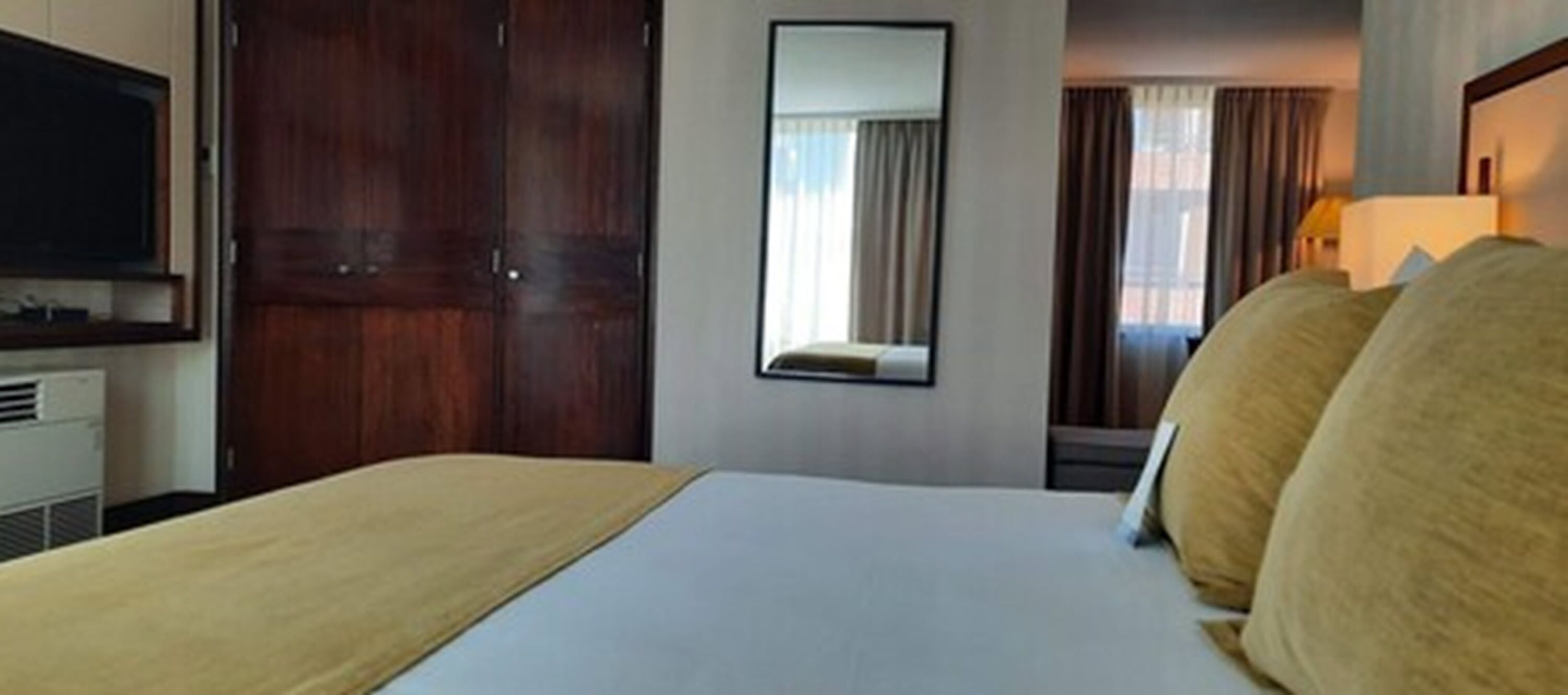 A hotel room with a bed, a TV, a mirror, and a window with curtains.