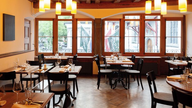 Un restaurant avec des tables et des chaises en bois, de grandes fenêtres et des lampes suspendues.
