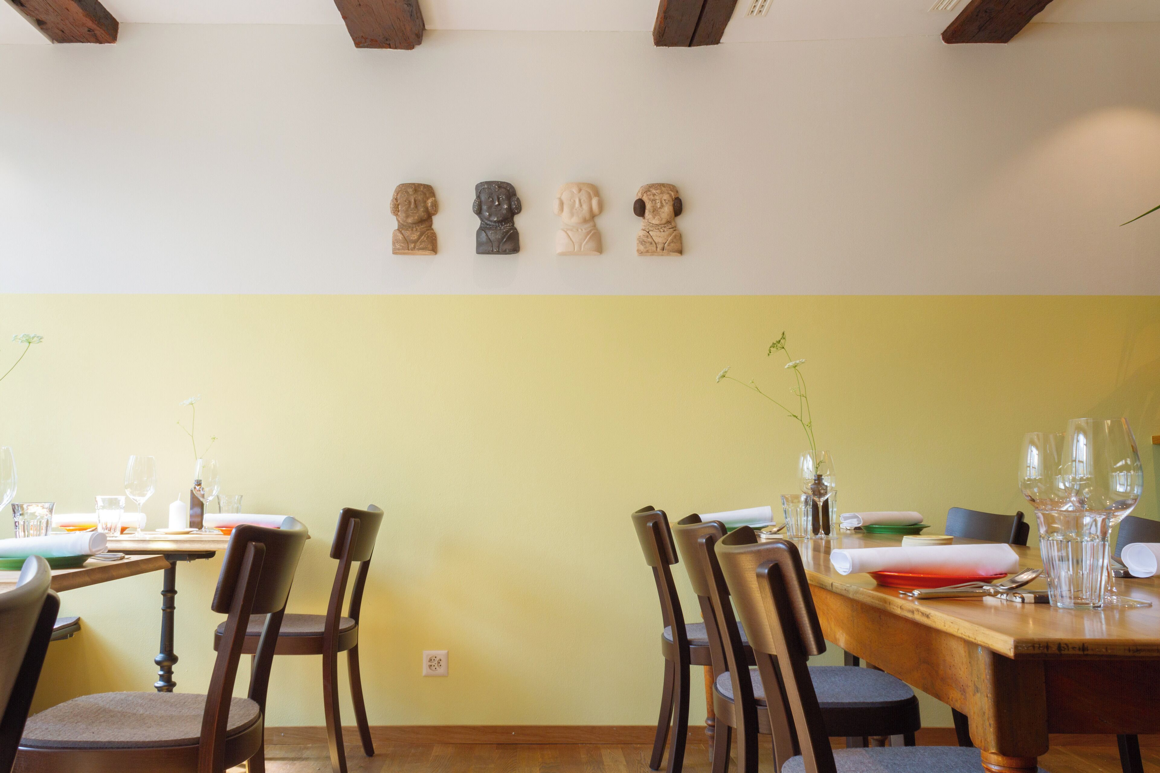 Un comedor con mesas y sillas de madera, una pared amarilla y cuatro esculturas de piedra.