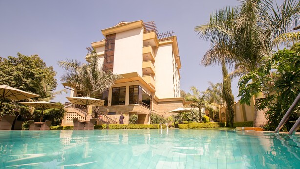 A swimming pool with lounge chairs and umbrellas, surrounded by palm trees and a multi-story building with balconies. - Waridi Paradise Hotel & Suites (Nairobi)
