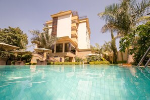 A swimming pool with lounge chairs and umbrellas, surrounded by palm trees and a multi-story building with balconies. - Waridi Paradise Hotel & Suites (Nairobi)