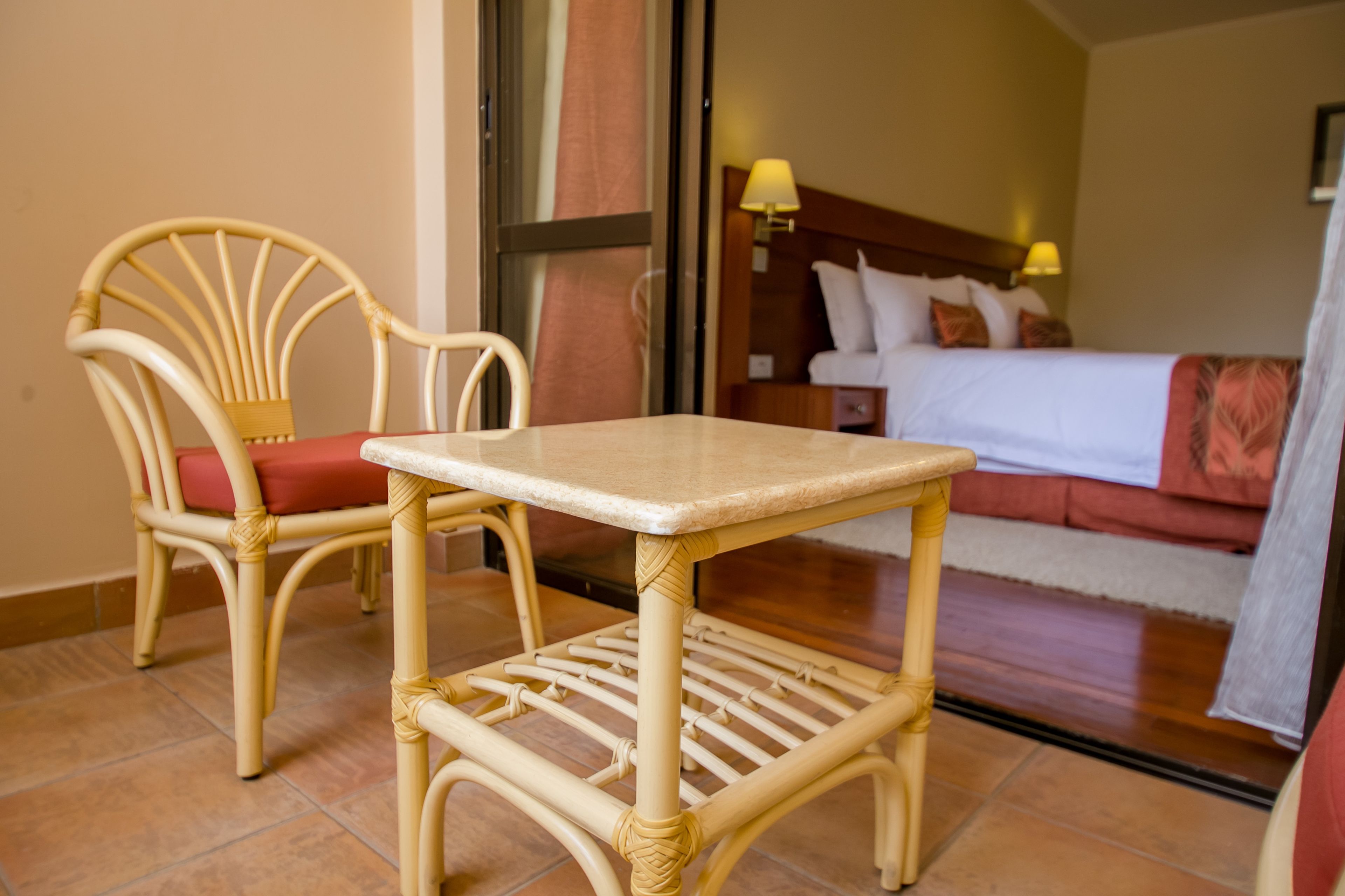 A hotel room with a bed, a nightstand, a lamp, a chair with a cushion, and a small table.