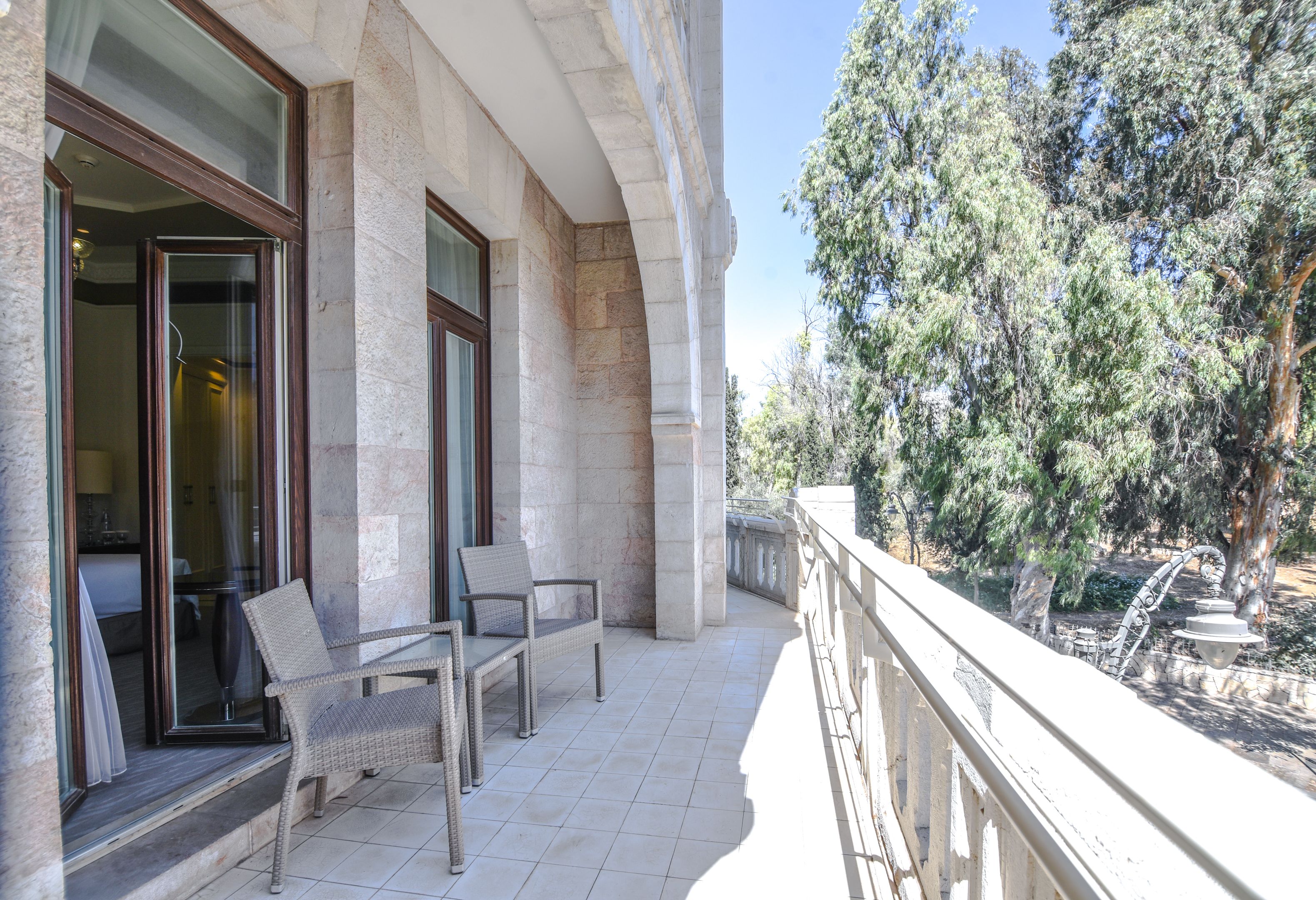 A balcony with wicker chairs and a table, overlooking a garden with trees and a fountain.