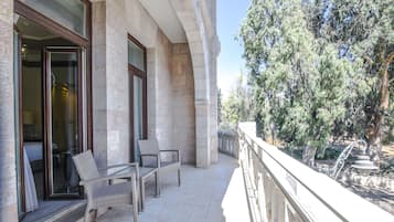 A balcony with wicker chairs and a table, overlooking a garden with trees and a fountain.