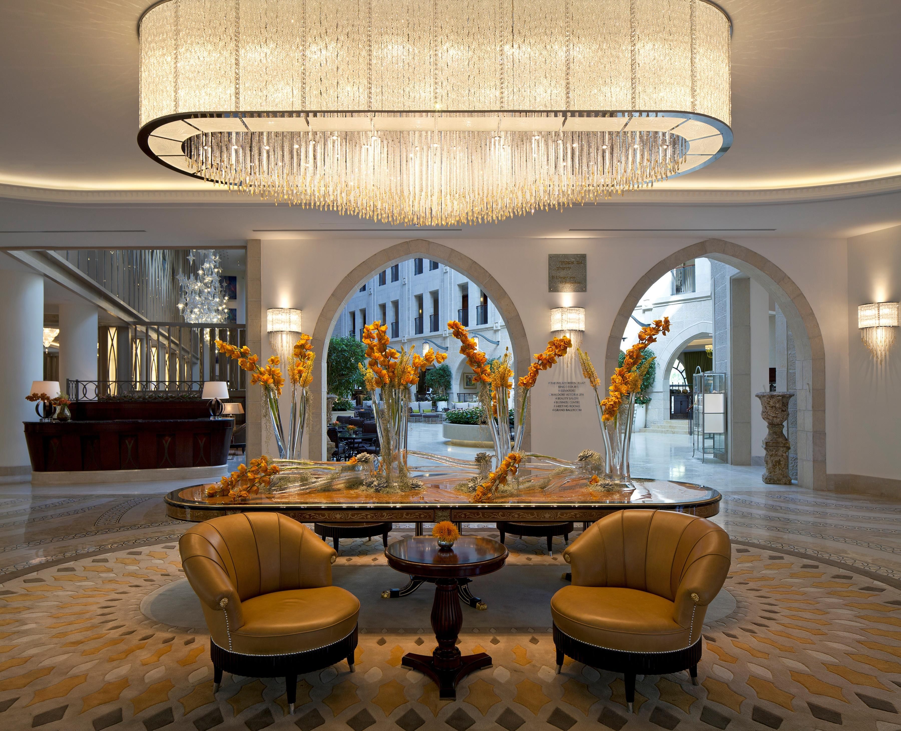 A lobby with a grand chandelier, two leather armchairs, a central table with floral arrangements, and a reception desk.