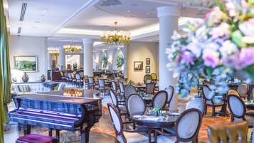 Uma ampla sala de jantar com várias mesas e cadeiras, um piano de cauda e lustres elegantes.