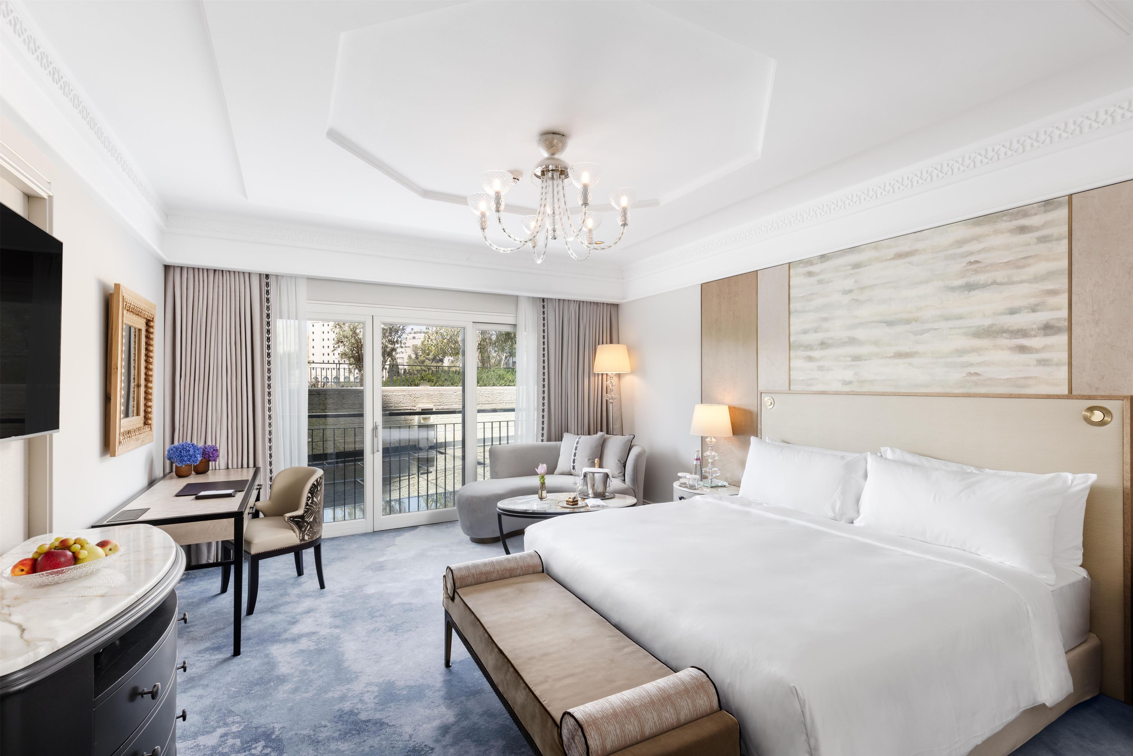Habitación de hotel con una cama grande, un escritorio, una silla, una mesita, un banco, un televisor y un balcón con vistas.