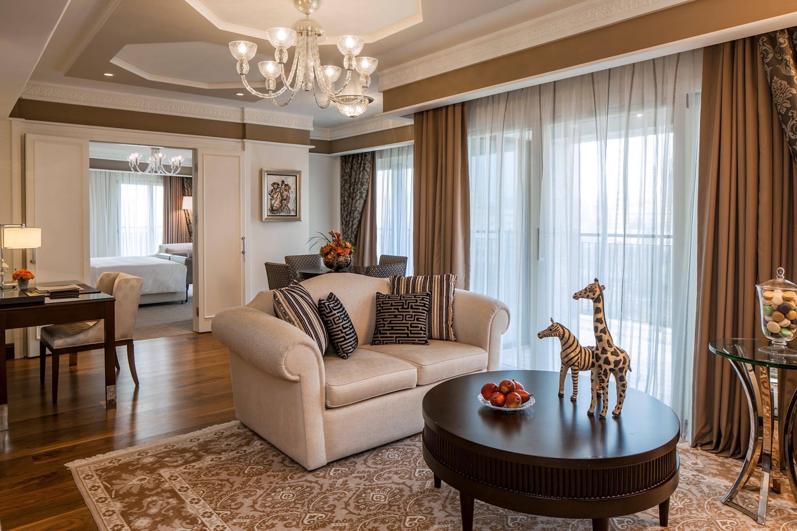 A well-lit living room with a sofa, a round coffee table, a chandelier, and a dining area with a table and chairs.
