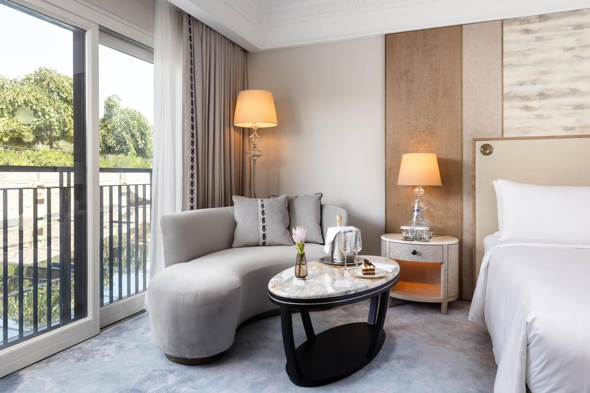 A hotel room with a sofa, a round table with a vase and a bottle, a nightstand with a lamp, and a bed.