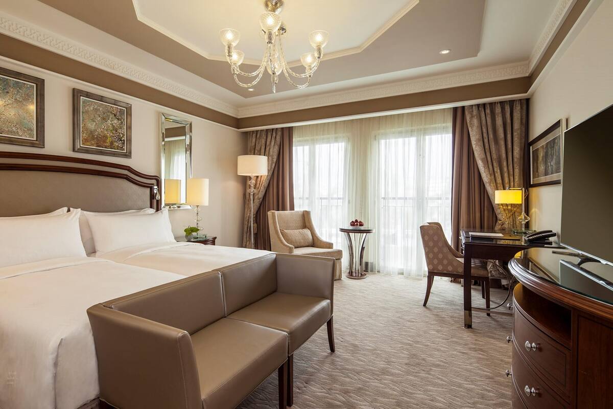 A hotel room with a large bed, a sofa, a desk, a chair, a TV, and a chandelier.