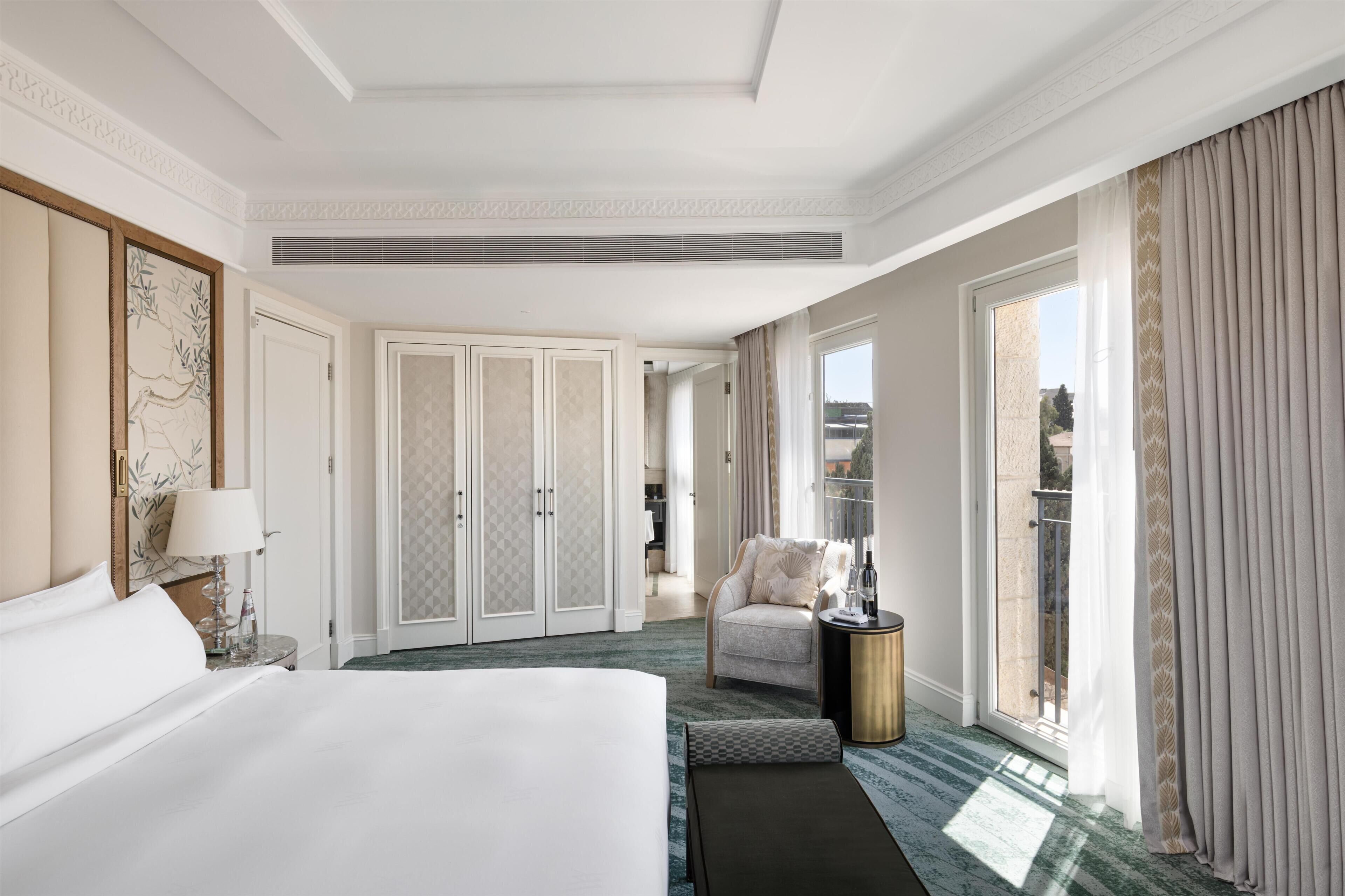 Habitación de hotel con una cama grande, una silla, una mesita, una ventana con cortinas y una puerta que da a un balcón.