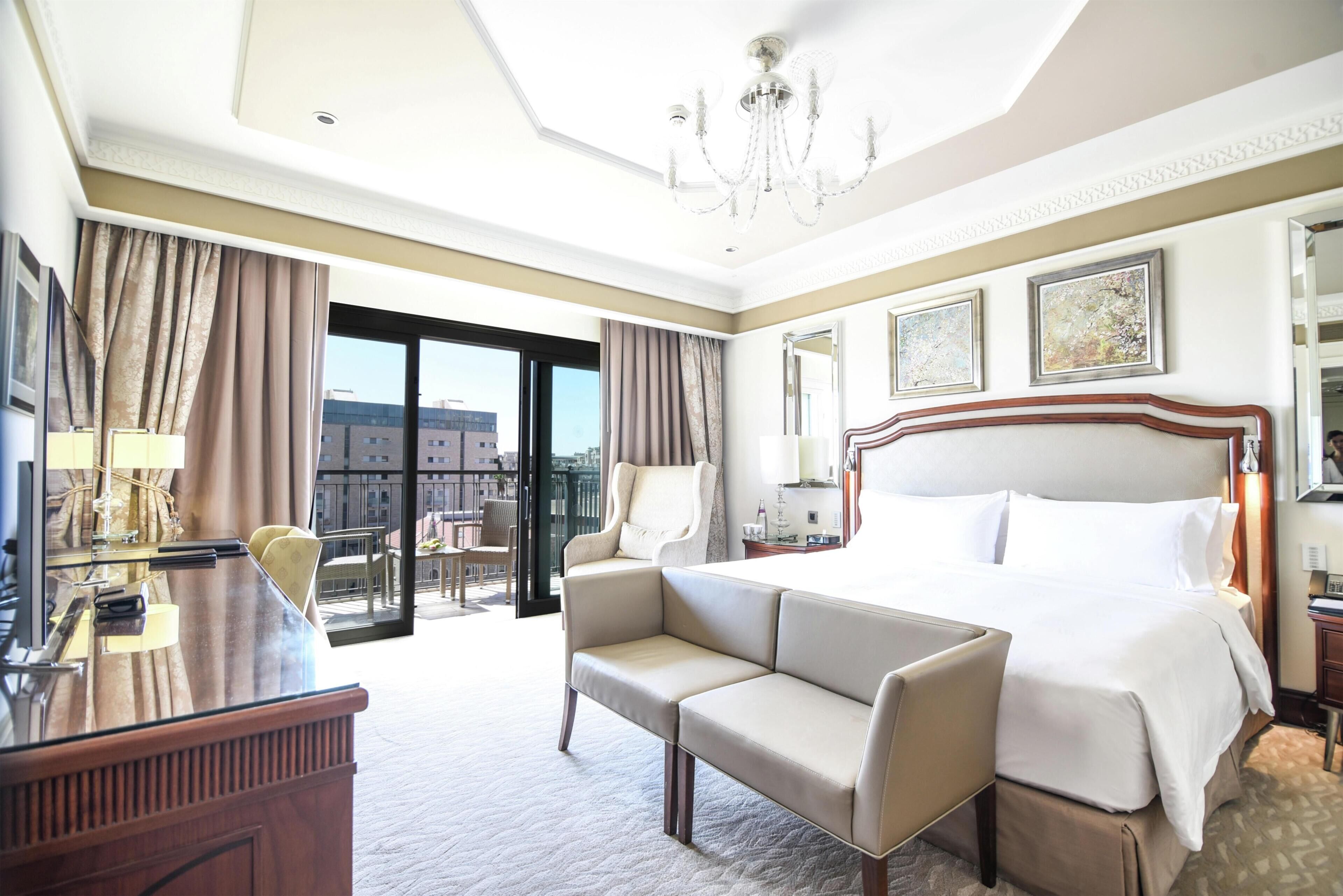 Habitación de hotel con cama, sofá, escritorio, silla, candelabro y vistas a edificios.