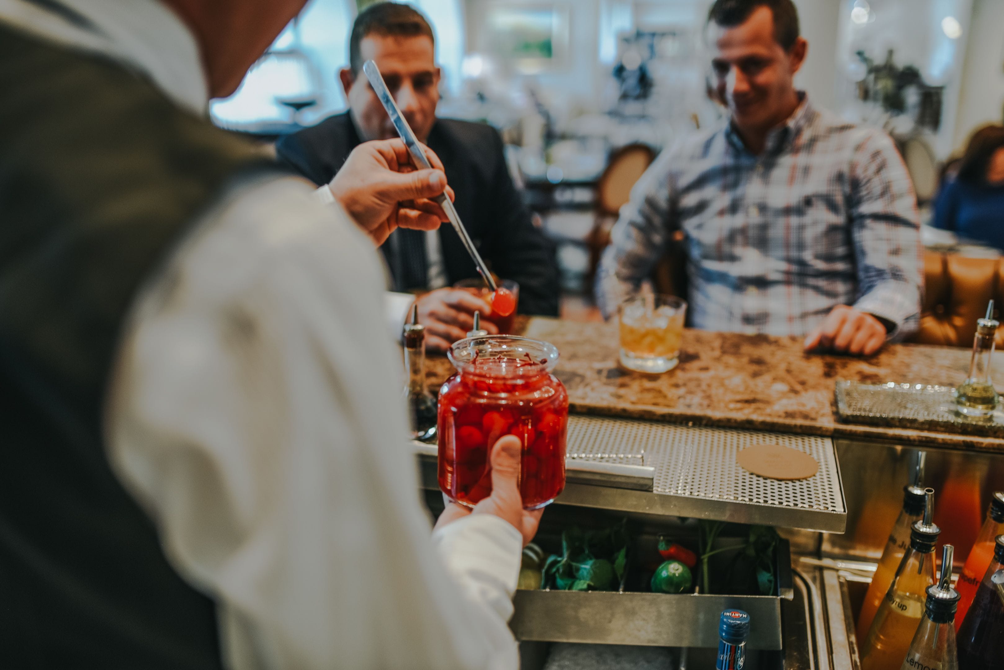 Zona de bar con una persona sosteniendo un tarro de bebida roja, un barman preparando una bebida y una barra con varias botellas.