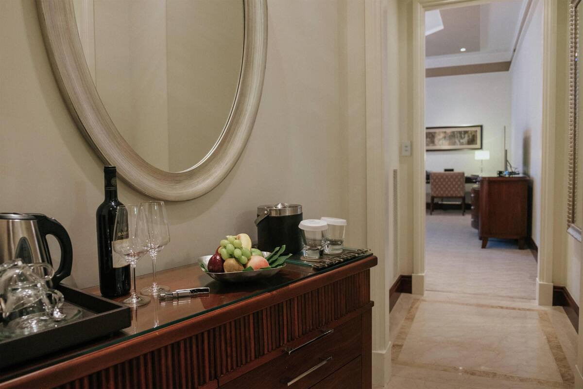 A hotel reception area with a wooden counter, a large mirror, a tray with a wine bottle, glasses, and a fruit bowl.