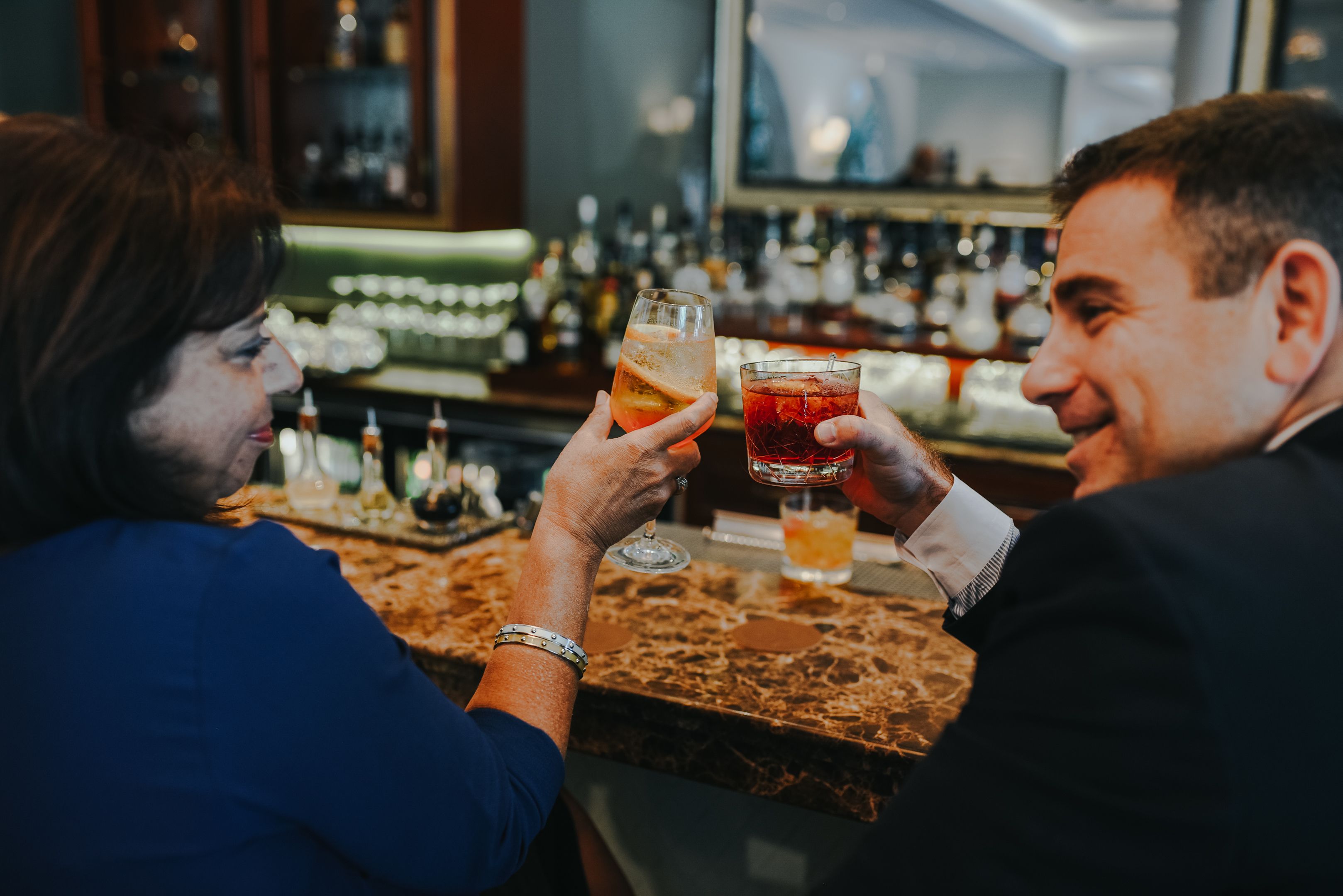 Duas pessoas brindando com bebidas em um bar com balcão de mármore e diversas garrafas ao fundo.