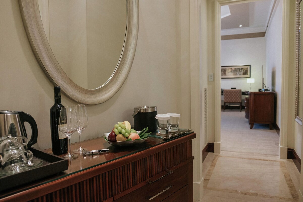 A hotel reception area with a wooden counter, a large mirror, a tray with a wine bottle, glasses, and a fruit bowl.