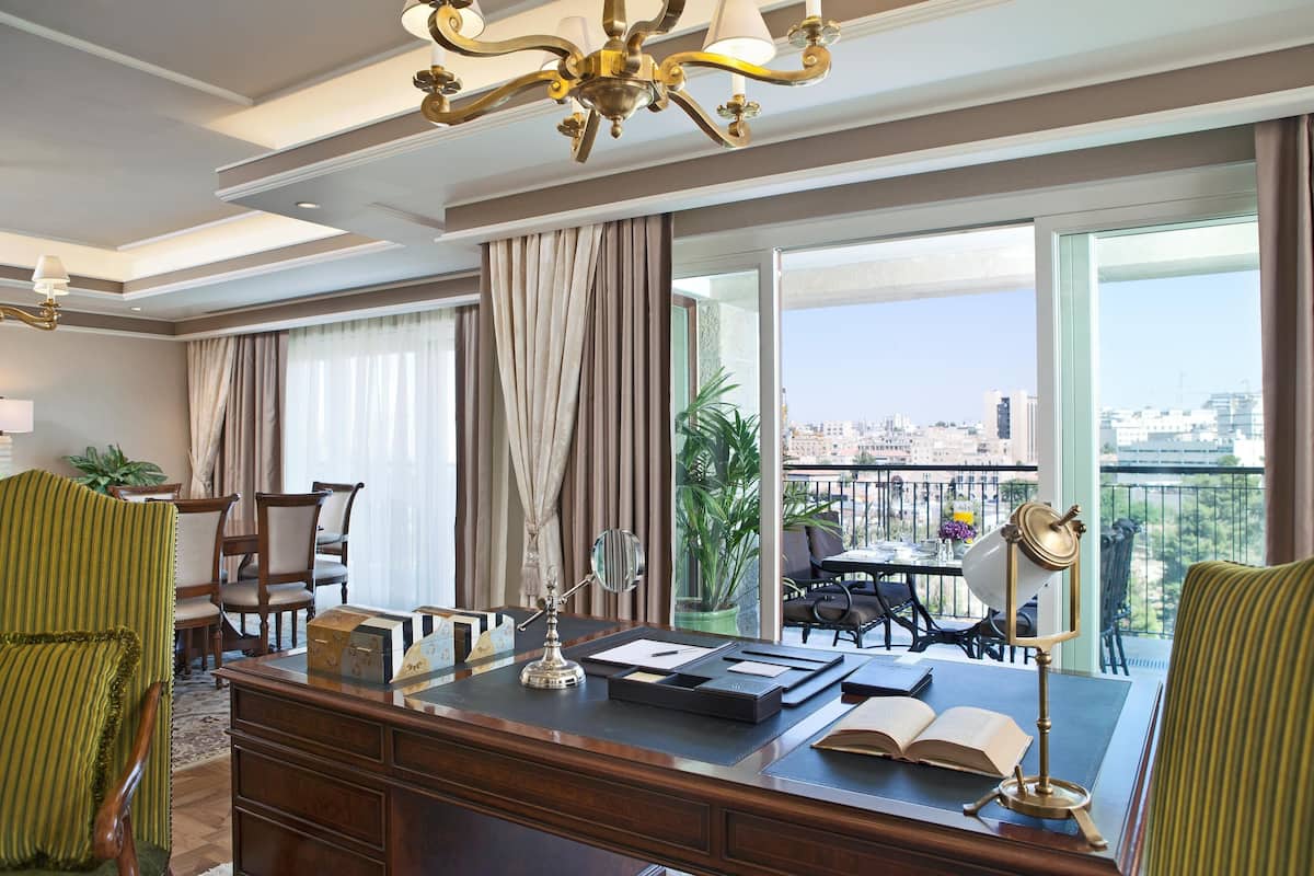 A hotel room with a desk, chairs, a chandelier, a large window with a city view, and a balcony with outdoor seating.