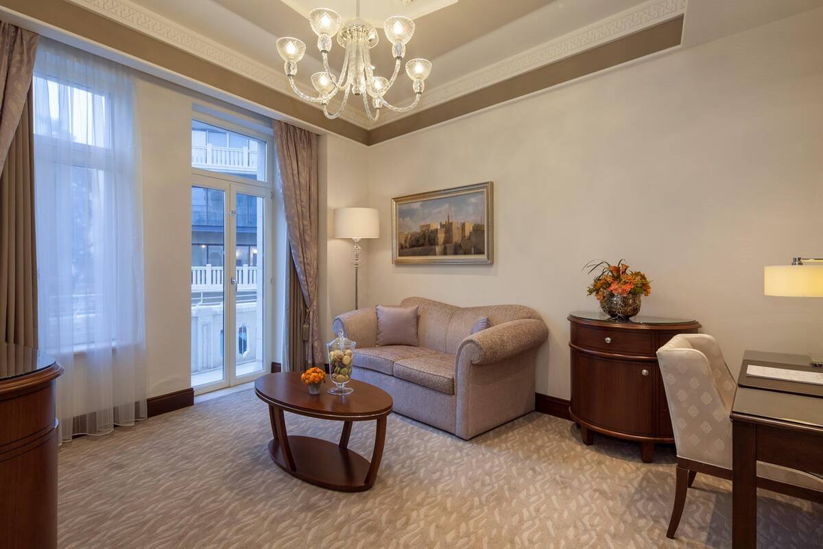 A hotel room with a sofa, a small table with flowers, a lamp, a painting on the wall, and a view through a window.