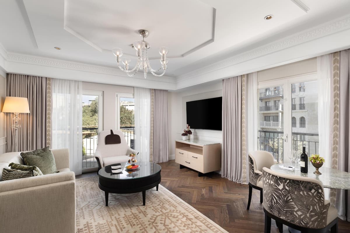 A spacious living room with a chandelier, a flat-screen TV, a dining area, and a balcony with a view.