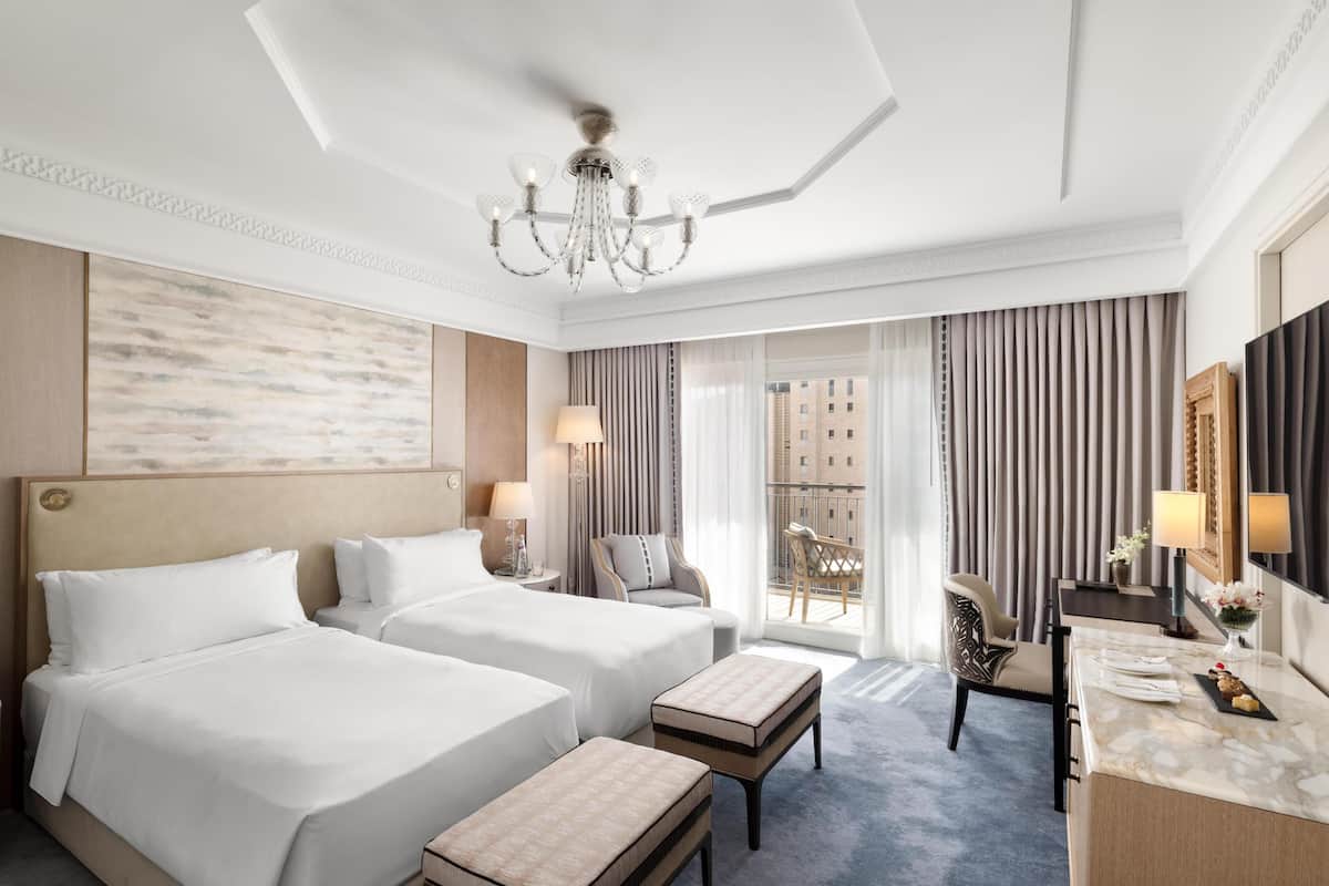 A hotel room with two beds, a chandelier, a desk with a mirror, and a view of buildings through large windows.