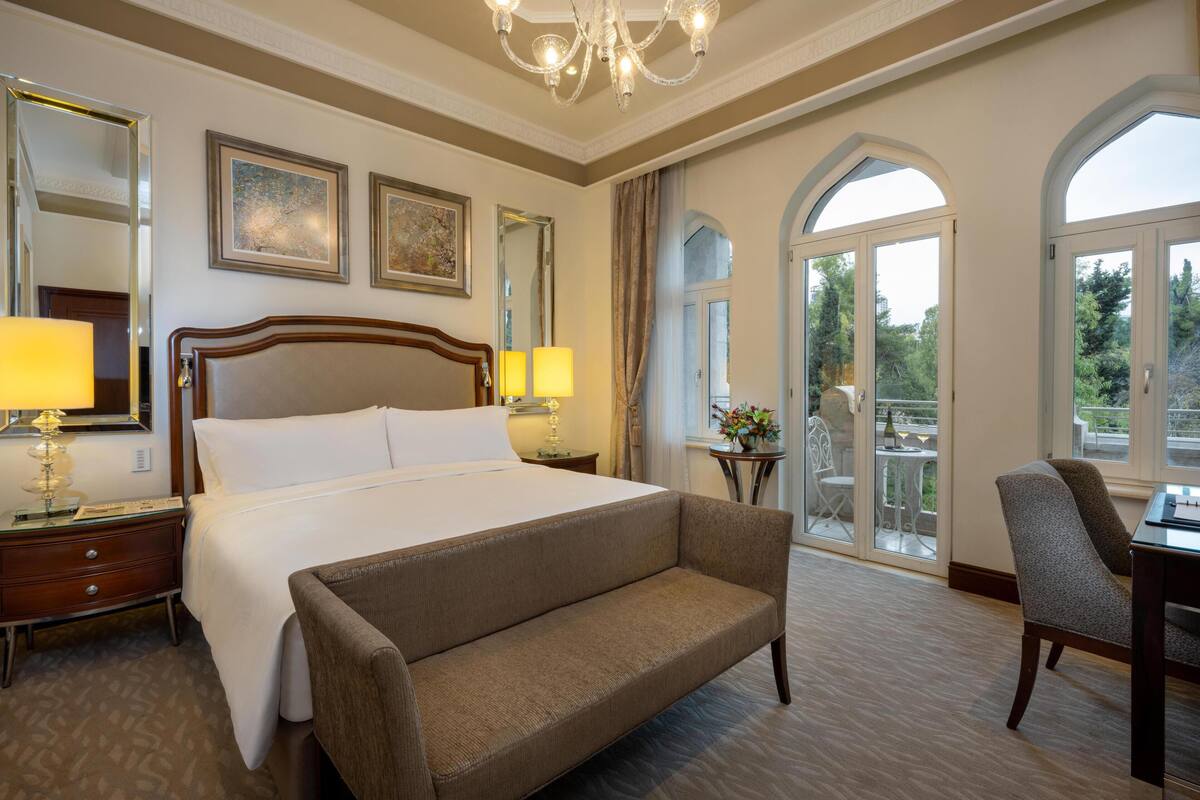 A hotel room with a large bed, two bedside lamps, a sofa, a desk, and a balcony with a view.