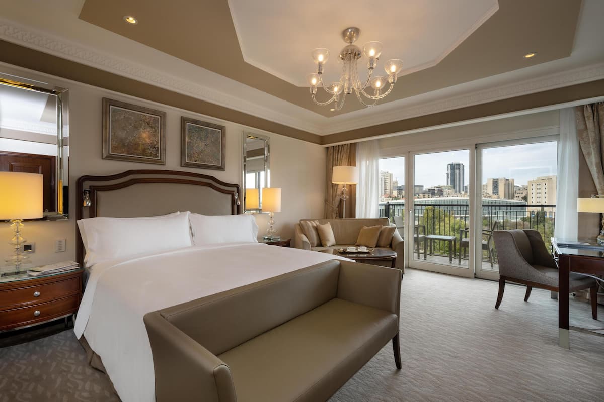 A hotel room with a large bed, a sofa, a desk, and a balcony with a city view.