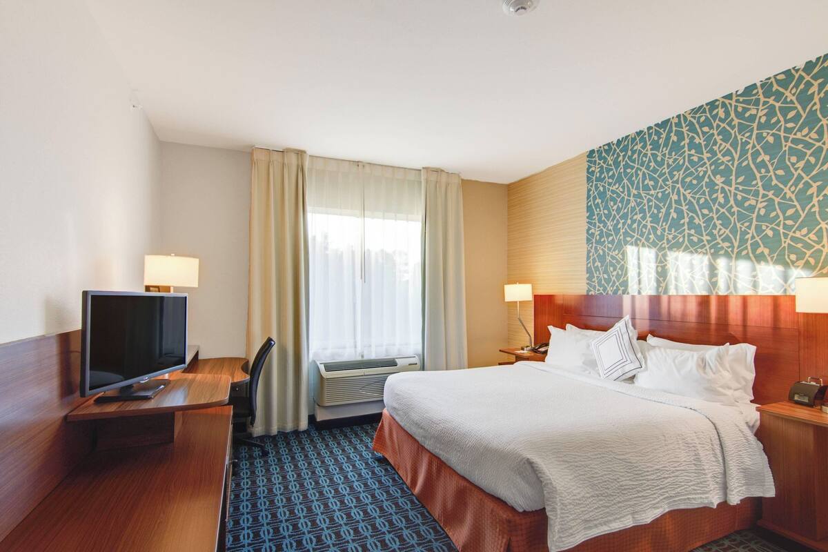 A hotel room with a bed, a desk with a TV, a window with curtains, and a patterned wall.