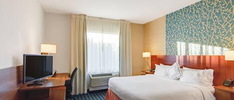 A hotel room with a bed, a desk with a TV, a window with curtains, and a patterned wall.