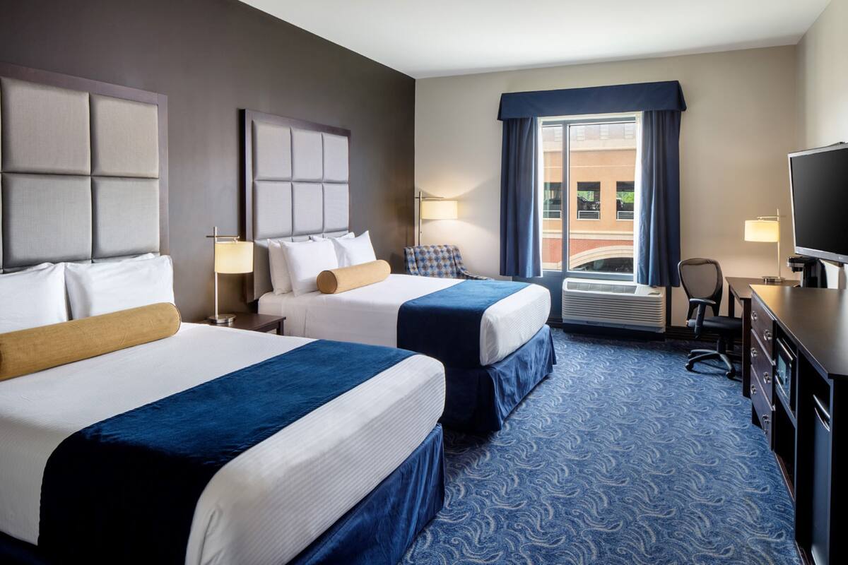 A hotel room with two beds, a large window with curtains, a desk with a television, and an air conditioning unit.