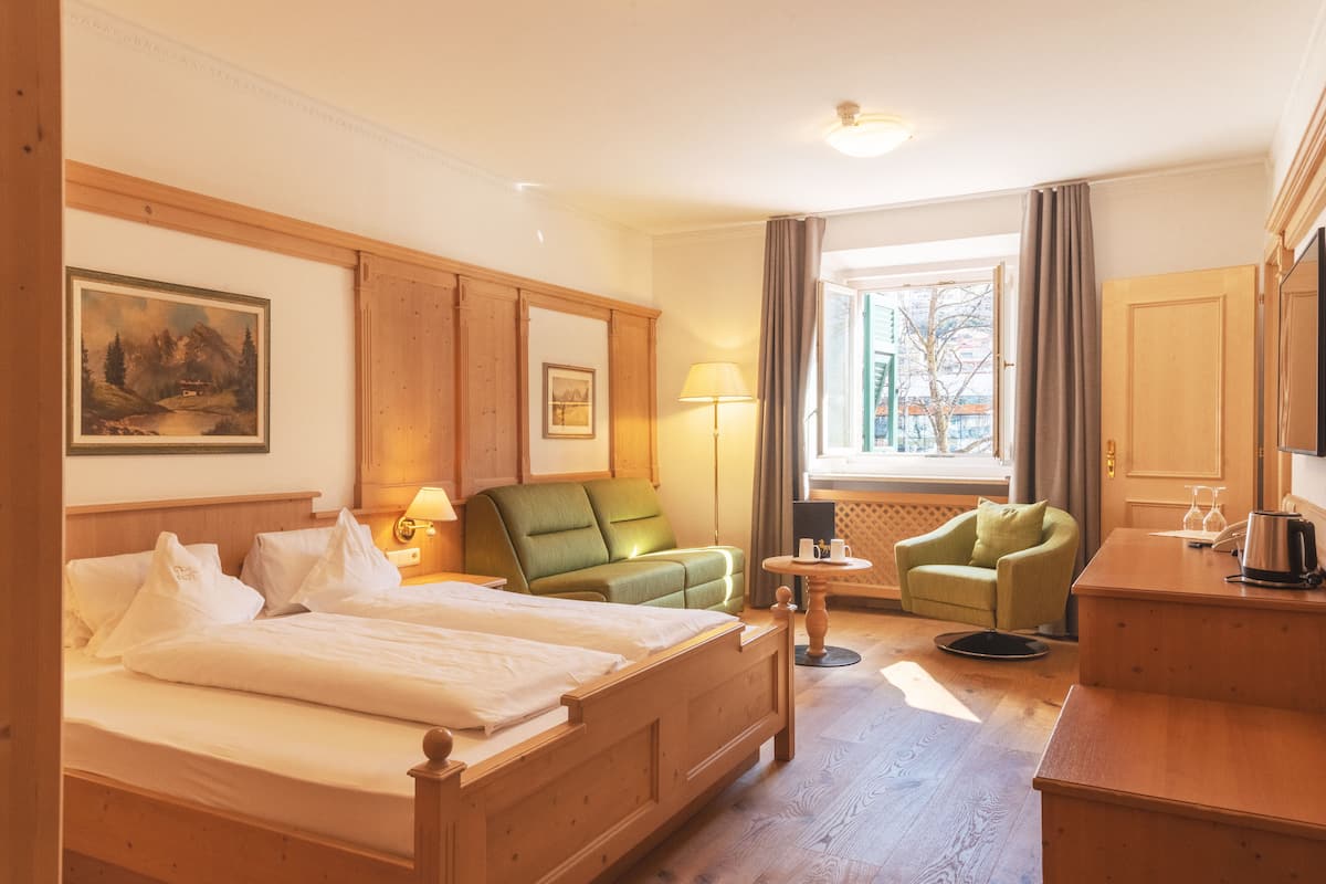 A hotel room with a wooden bed, a green armchair, a small round table, and a view of the outside through a window.