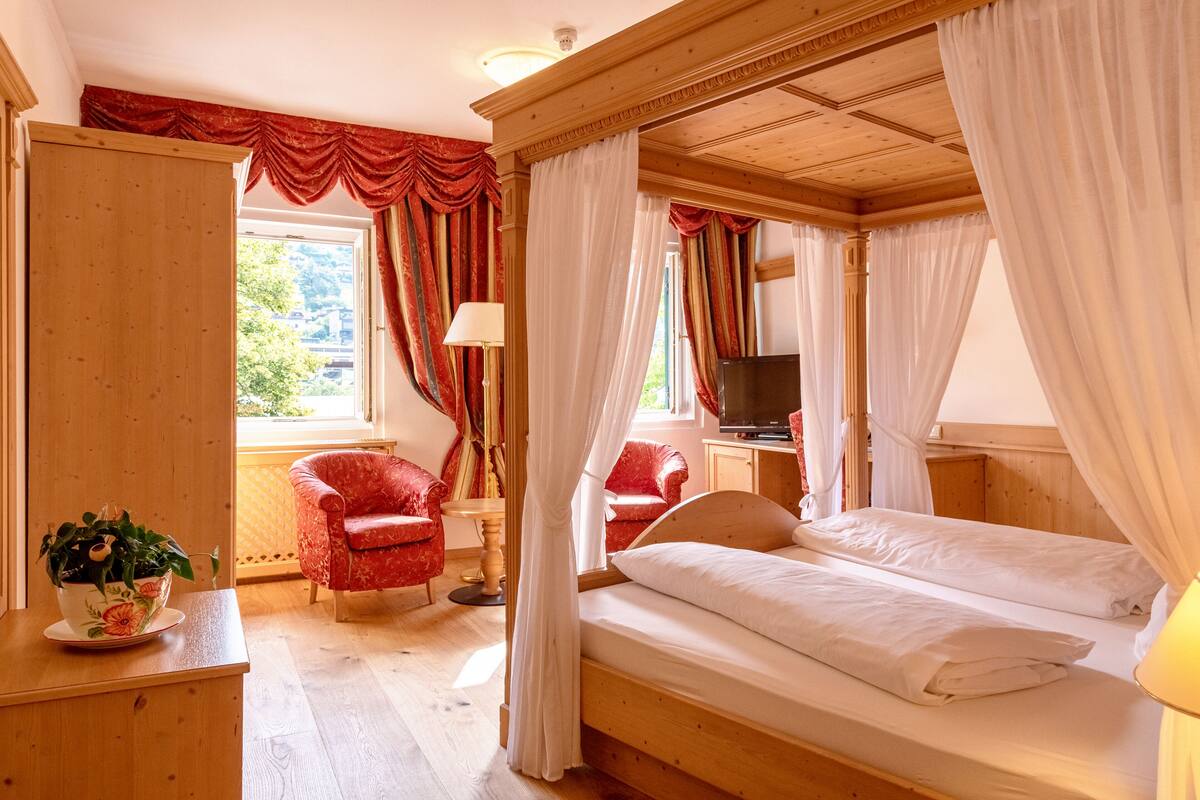 A wooden four-poster bed with white linens, a red armchair, a small table with a plant, and a television on a wooden cabinet.