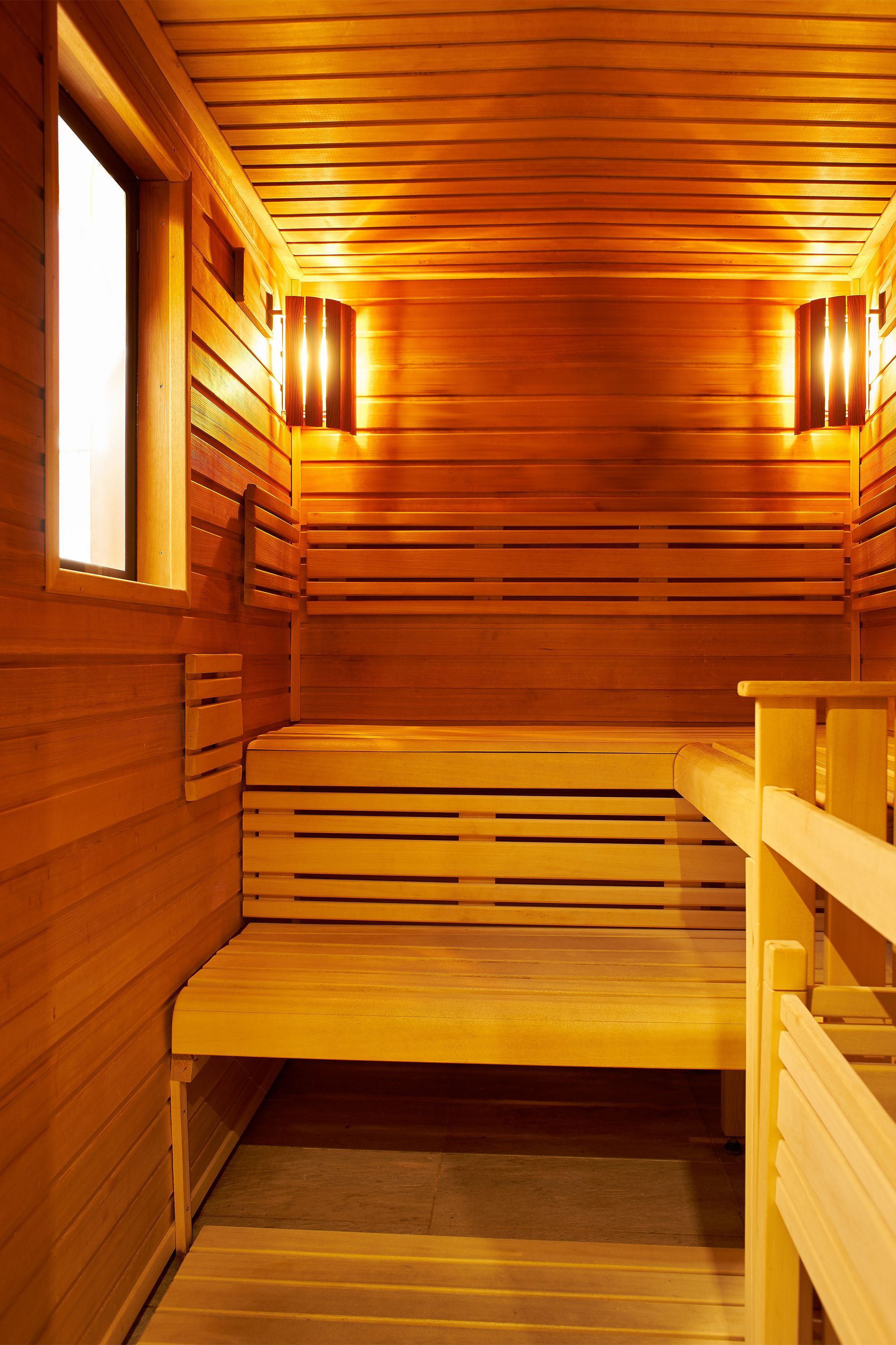 Uma sauna com bancos e paredes de madeira, uma janela e luminárias fixadas na parede.