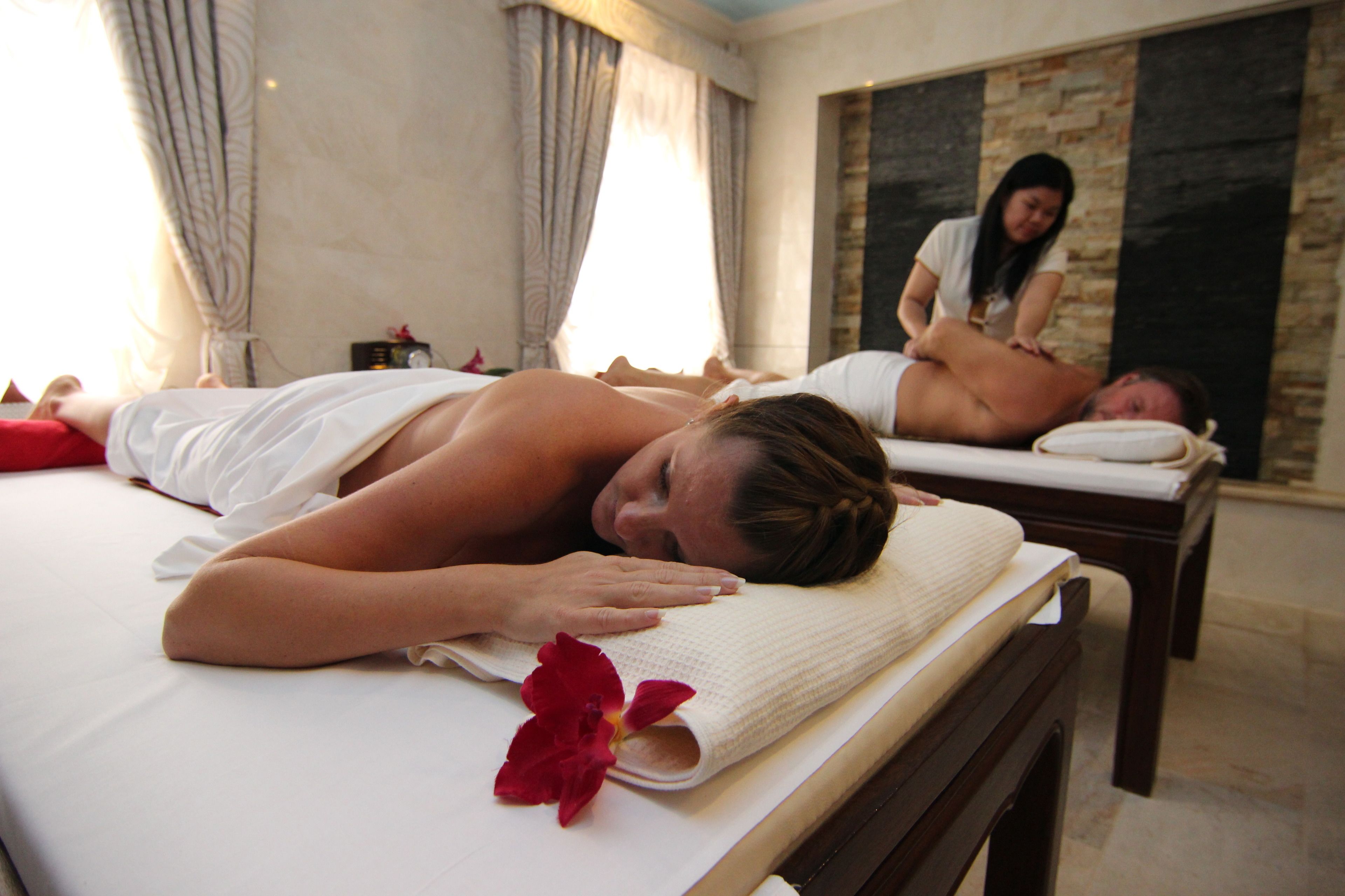 A spa room with two massage tables, one occupied by a person receiving a massage and another with a person lying down. A massage therapist is attending to the person on the table.