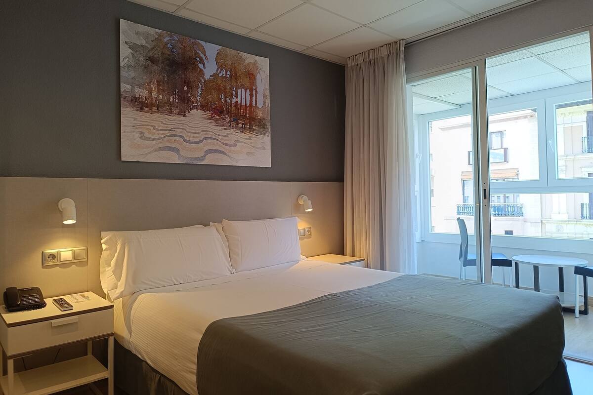 Une chambre d’hôtel avec un grand lit, une table de chevet avec un téléphone et un livre, un tableau au mur et une vue sur l’extérieur par une fenêtre.