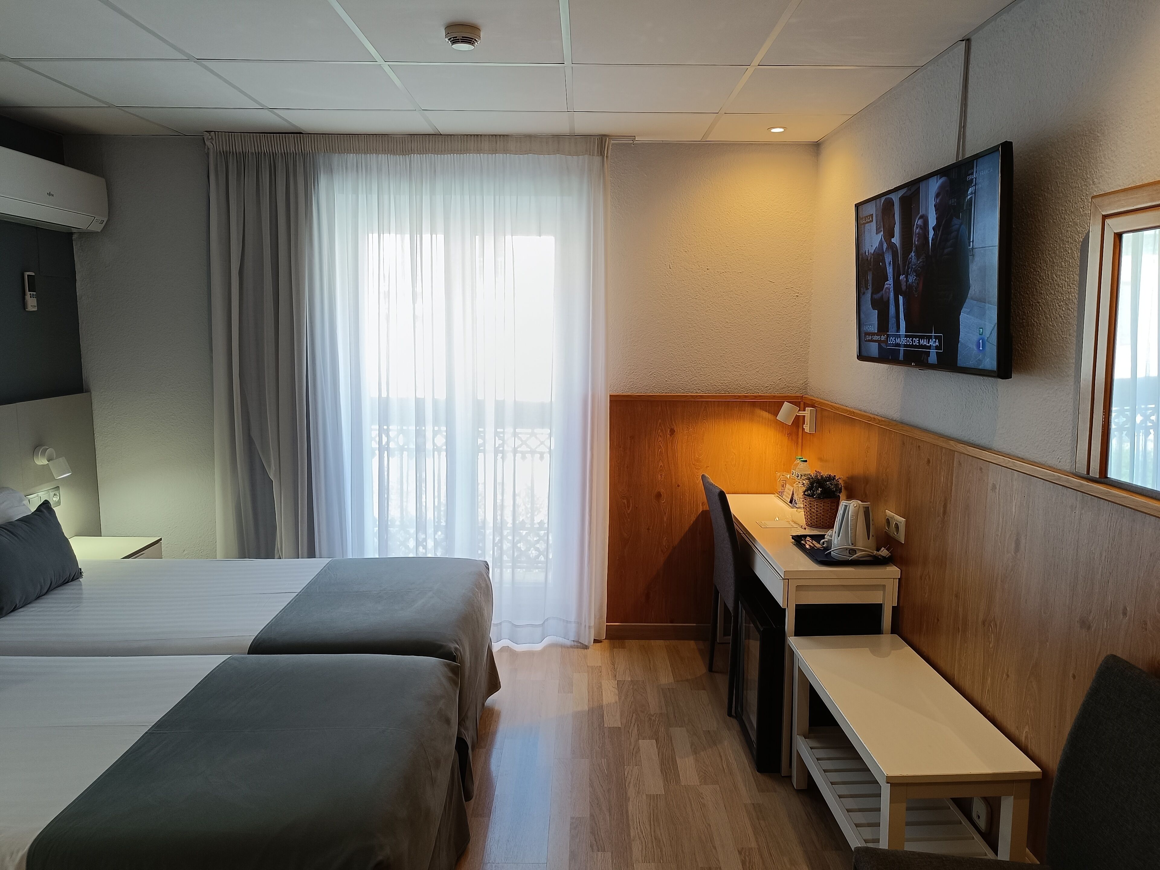 A hotel room with two beds, a desk with a chair, a TV mounted on the wall, and a balcony with a railing.