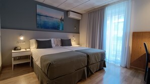 A hotel room with a double bed, bedside tables, a nightstand, a wall-mounted air conditioner, and a large window with curtains.
