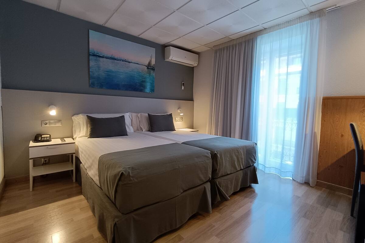Une chambre d’hôtel avec un lit double, des tables de chevet, une petite table, un climatiseur fixé au mur et une grande fenêtre avec des rideaux.