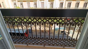 A balcony with a metal lattice railing, a tiled floor, and a view of a street with buildings and parked vehicles.