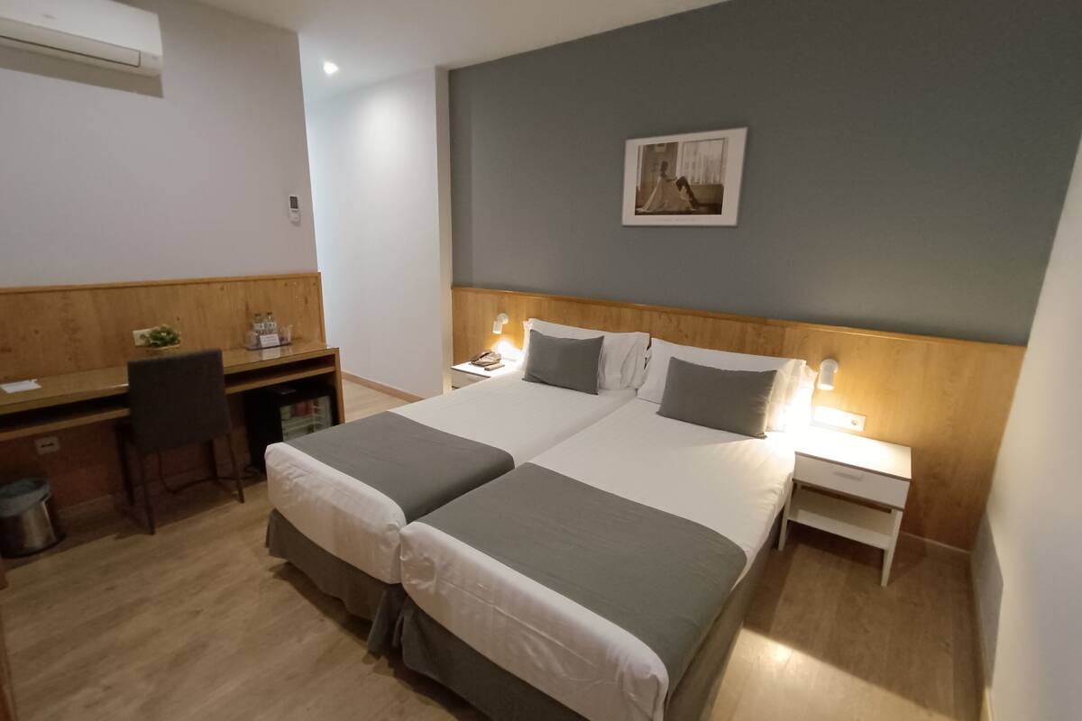 Une chambre d’hôtel avec un lit double, un bureau avec une chaise, une petite poubelle et un tableau accroché au mur.