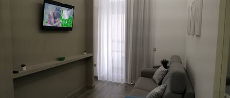 Una sala de estar con una televisión de pantalla plana montada en la pared, un sofá, una mesita con un cuenco y una planta, y una ventana con cortinas translúcidas.